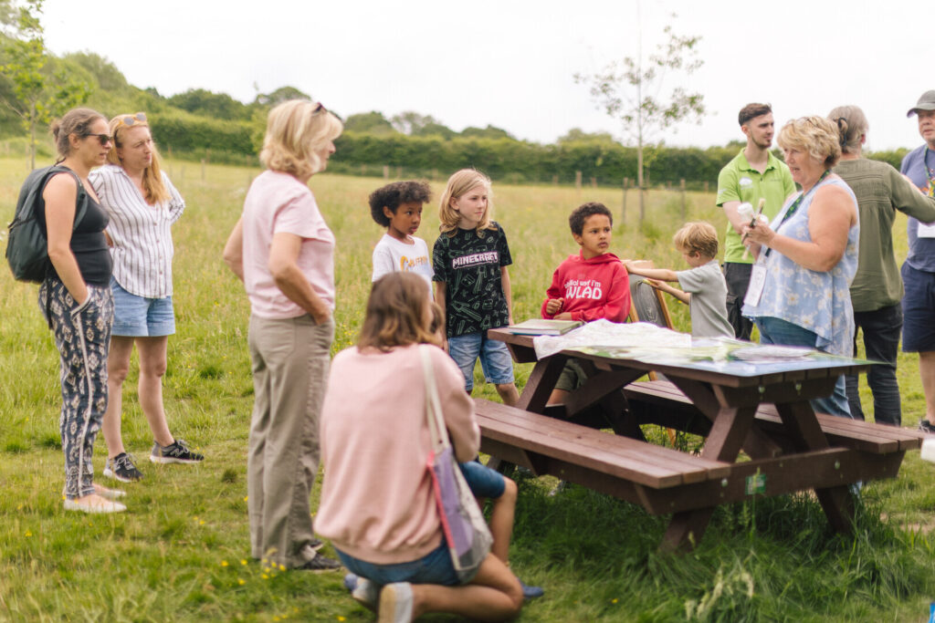 Group of adults and children gathered by a picnic table in a grassy field, listening to a guide explain maps and materials