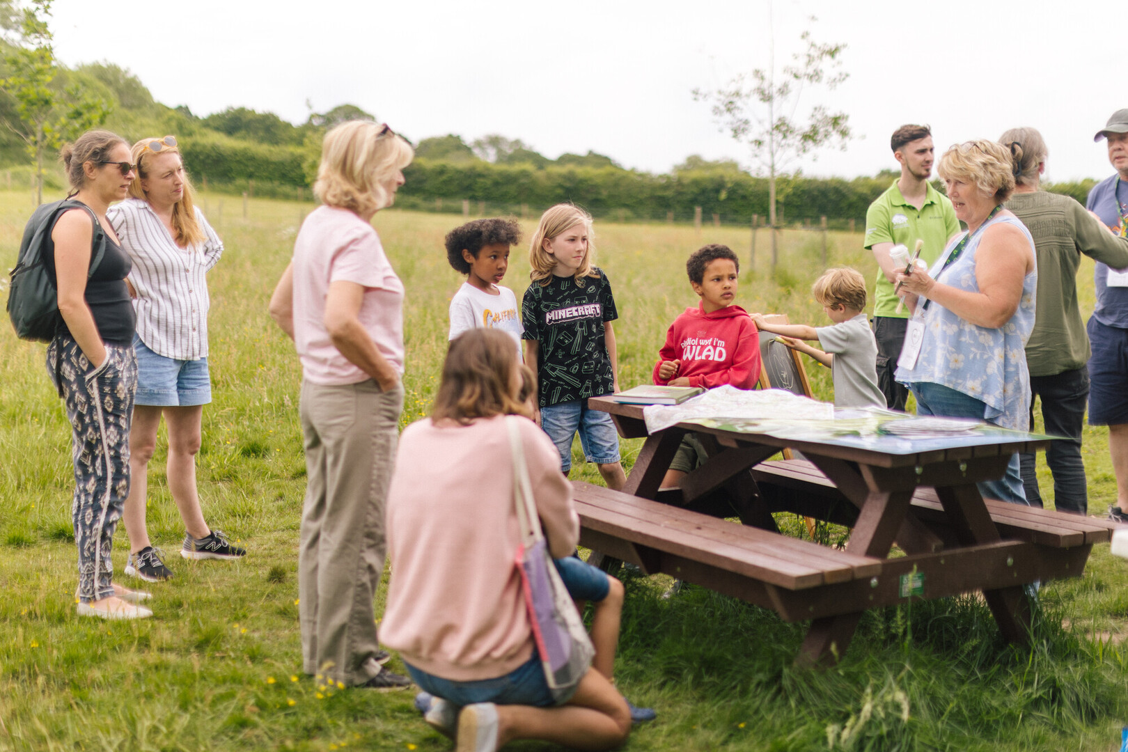 Group of adults and children gathered by a picnic table in a grassy field, listening to a guide explain maps and materials