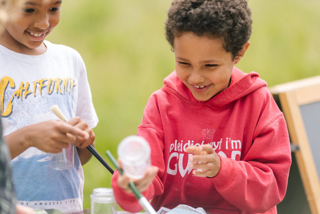 Two children smiling while painting outdoors, holding paintbrushes and clear plastic cups on a table