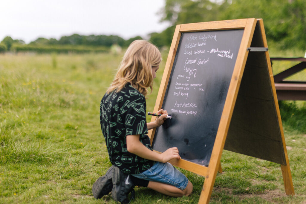 Child kneeling on grass writes on a chalkboard listing insects and butterflies during an outdoor nature activity