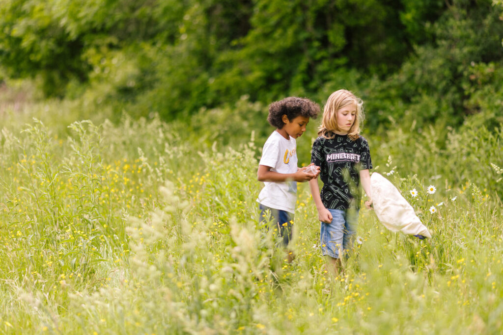 Two children stand in a wildflower meadow, one holding a net while the other examines something in their hands