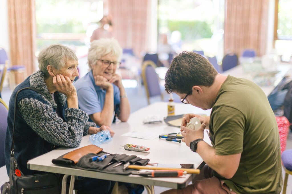 Man repairs a small white device at a community repair cafe table while two older women watch, tools and glue on the tabletop
