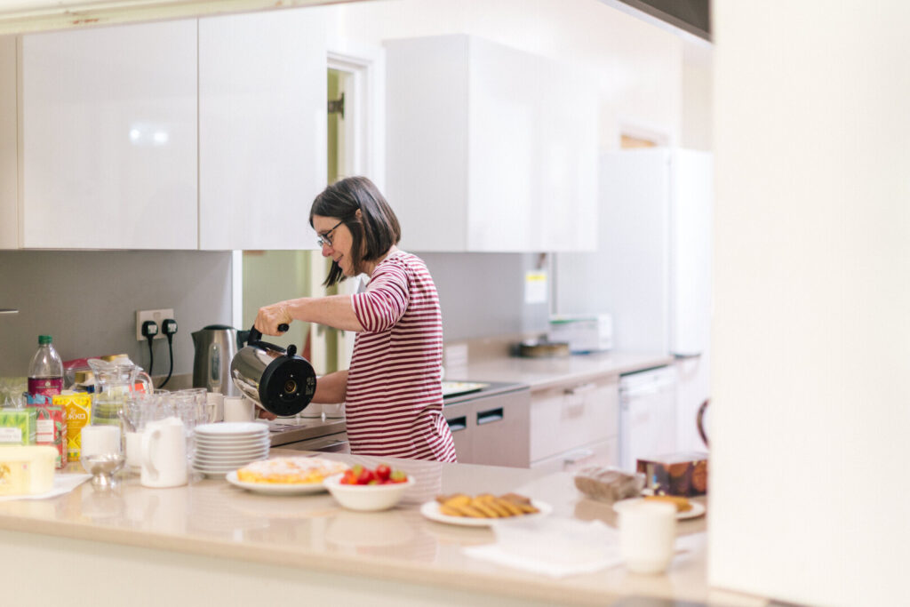 Woman pours hot water from a kettle in a community kitchen, with cups, tea supplies, and snacks on the counter