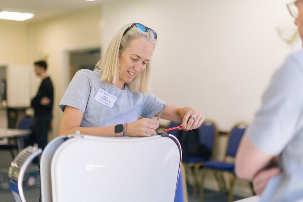 Woman at a repair café uses a screwdriver to fix an appliance while talking to someone across the table