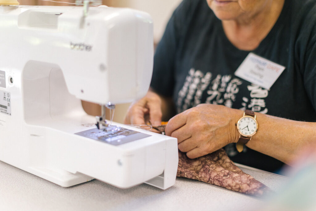 Person using a sewing machine to repair patterned fabric at a table, wearing a name badge and wristwatch
