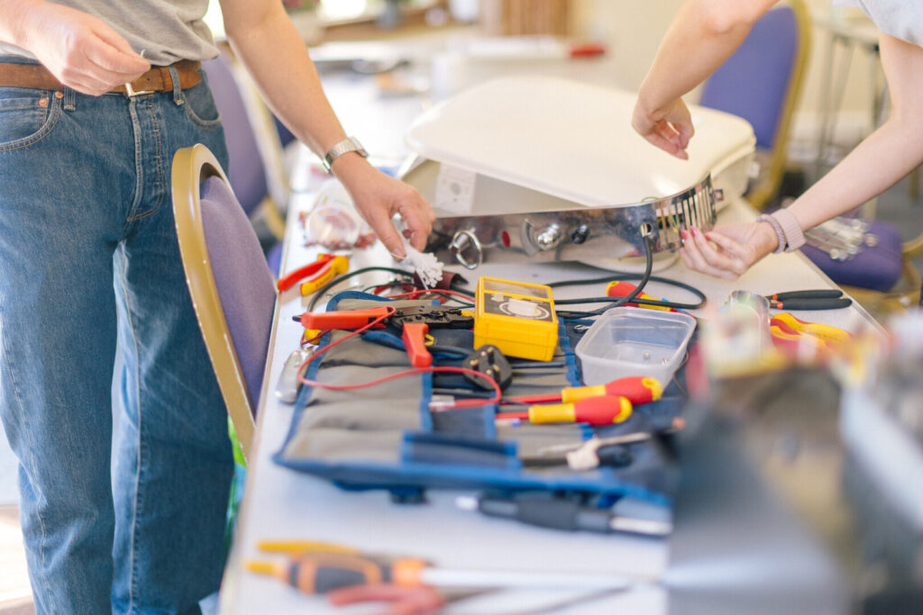Two people repair a kitchen appliance on a table with multimeter, wires, screwdrivers and pliers at a repair cafe