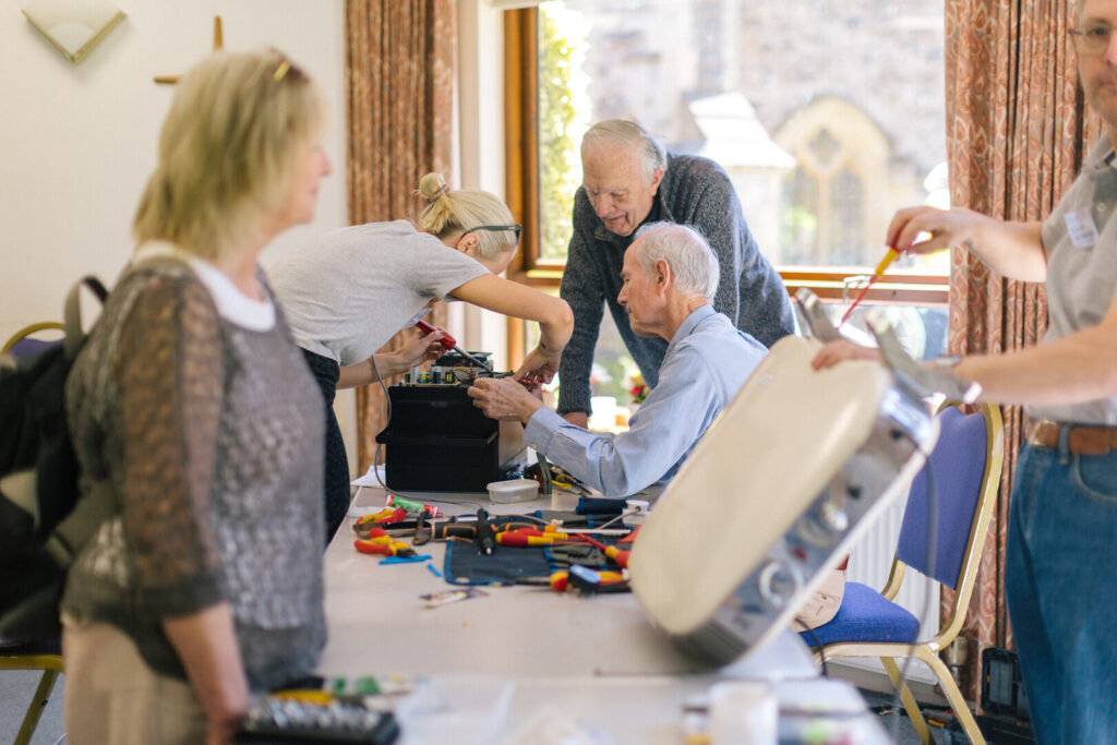 Volunteers and older men repair electronics at a community repair cafe table covered with tools and wires