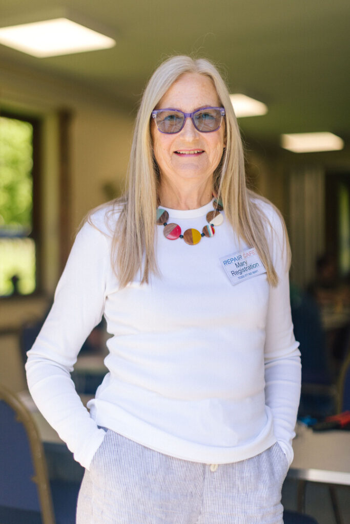 A woman with a white long sleeve t shirt, blue shades and a large necklace, stood in a church hall, she is smiling