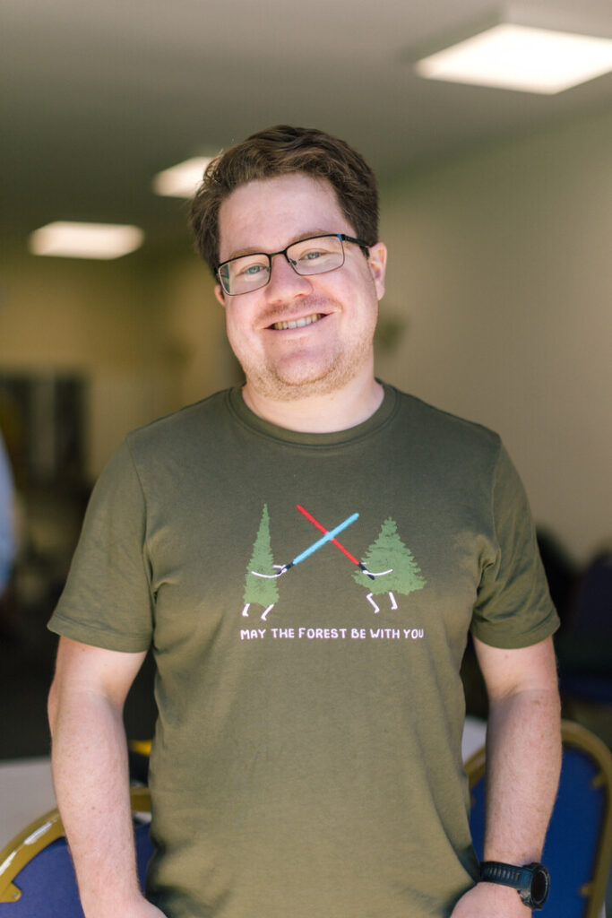 A man in a green t shirt stood in a church hall, he is smiling 