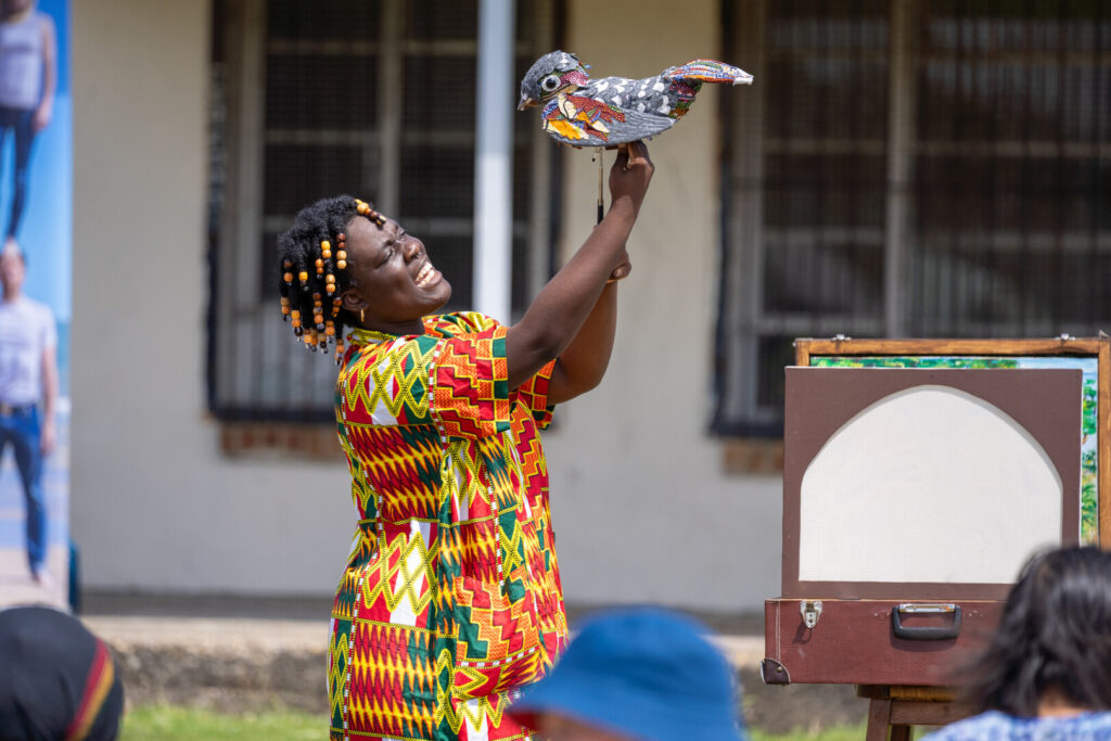 A person in bright clothing symbolic of Ghana holds a model nightjar up high and sings.