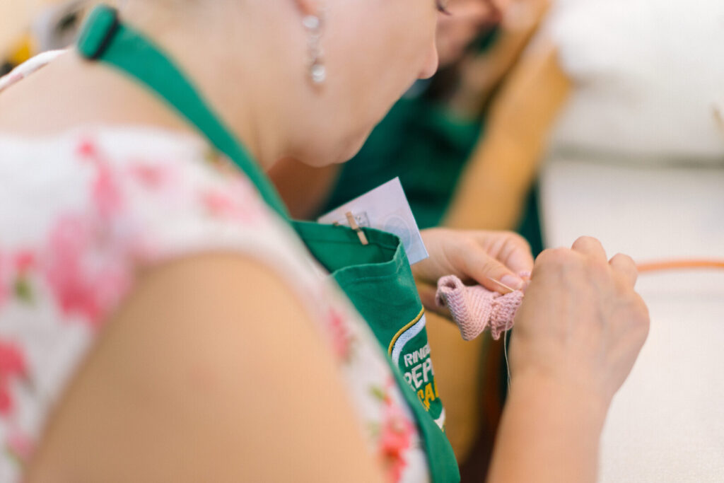 Person in green Ringwood Repair Cafe apron sewing a small pink knitted item at a table
