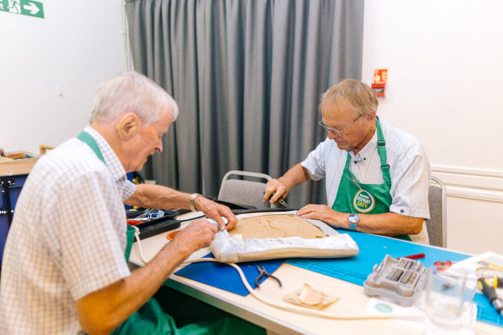 Two volunteers in green Repair Cafe aprons fix a chair seat at a worktable with tools and parts