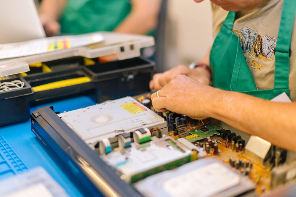 Person in green apron repairing an open DVD player, working on the circuit board at a workbench with toolboxes