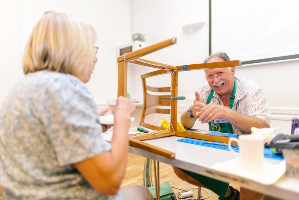Man in green apron repairs an upside-down wooden chair with clamps and string at a table, smiling at a seated woman