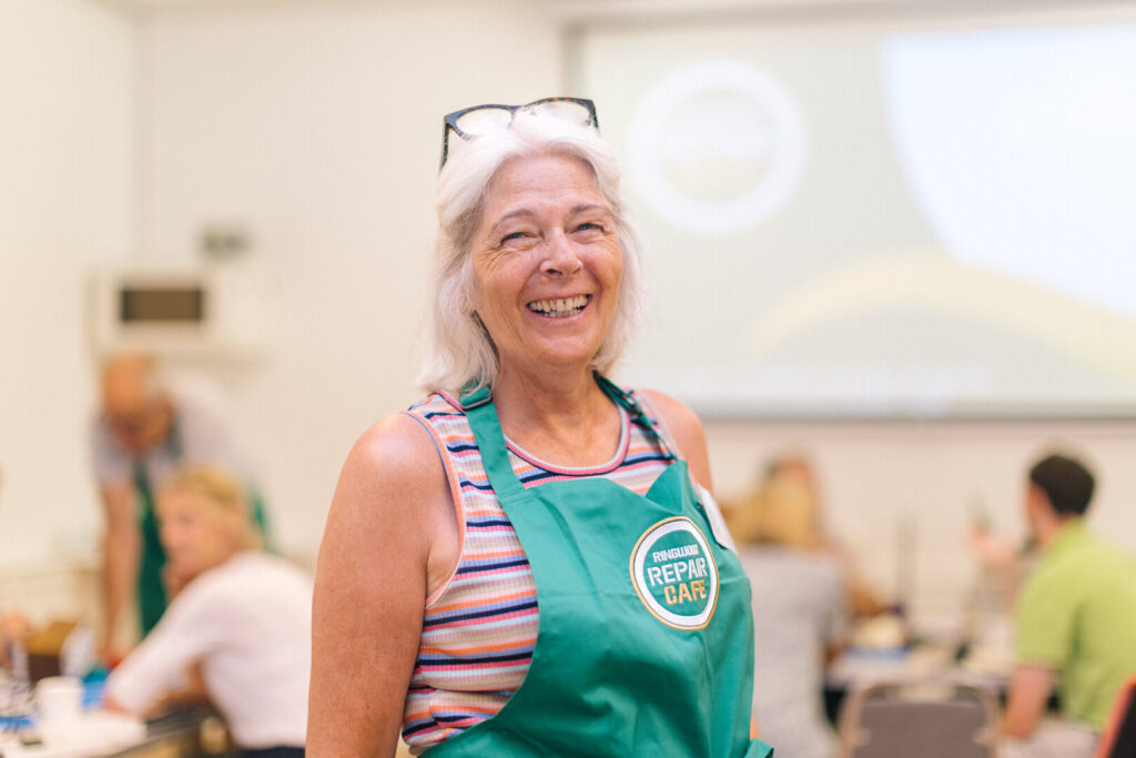 A woman with a green apron embroidered Ringwood Repair Cafe stood in a hall and smiling