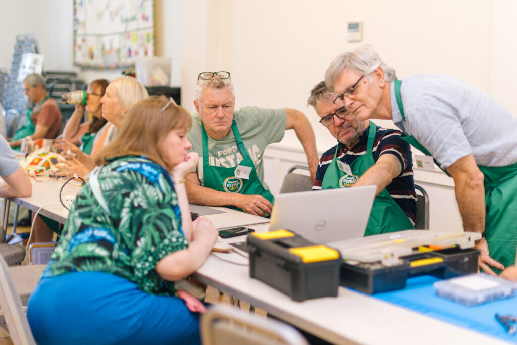 Volunteers in green aprons at Ringwood Repair Cafe help a woman with a laptop at a long table with tools and equipment