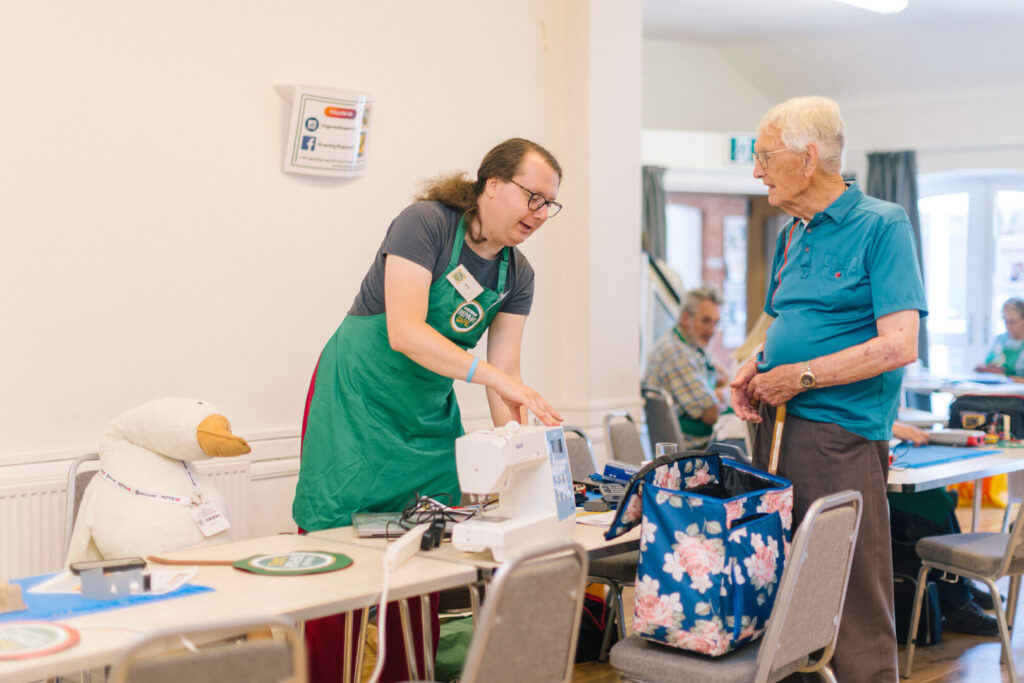 Volunteer in green apron demonstrates a sewing machine to an older man in a community repair cafe, with tools on the table