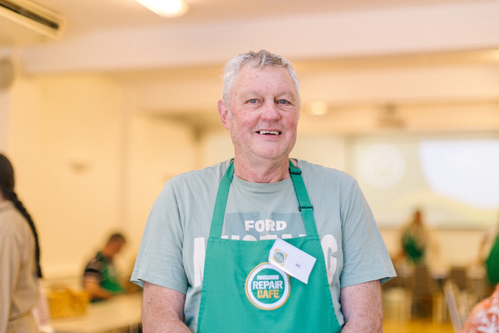 A man in a green Ringwood Repair Cafe apron stood in a hall and smiling
