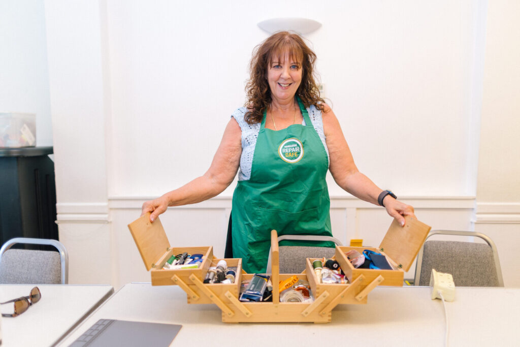 Woman in green Ringwood Repair Cafe apron stands behind table with open wooden toolbox of sewing and repair supplies