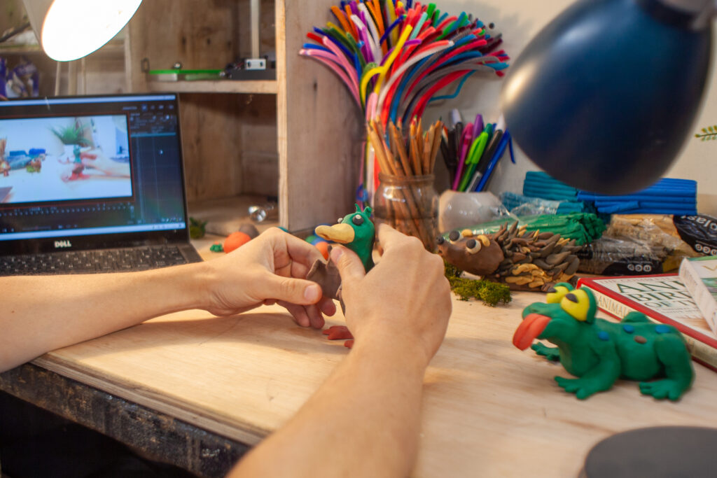 Hands sculpting a clay duck at a desk with art tools, clay animals, and a laptop showing a stop-motion scene