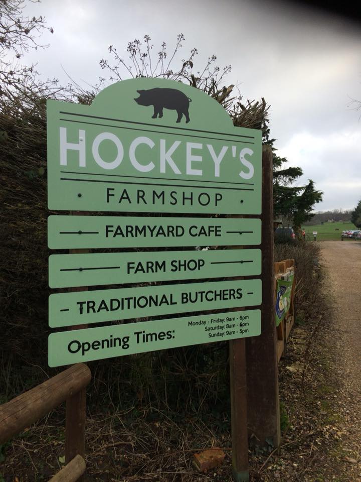 Green roadside sign for Hockey's Farmshop with pig silhouette, listing Farmyard Cafe, Farm Shop, Traditional Butchers and opening times