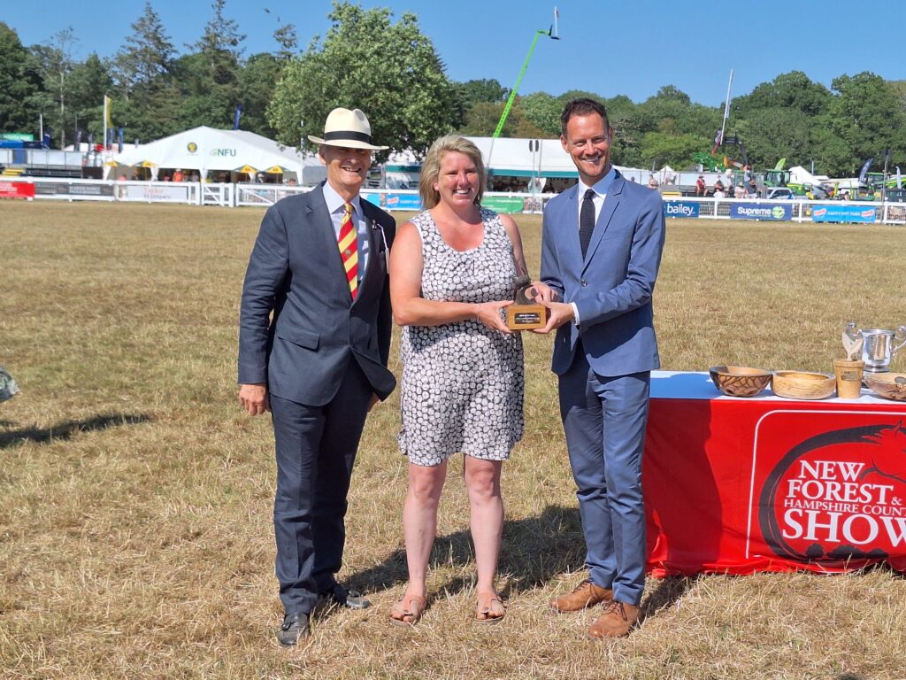 Elycia Farnish receives an award from two men at the New Forest Show 2026, standing by a table of trophies on a field
