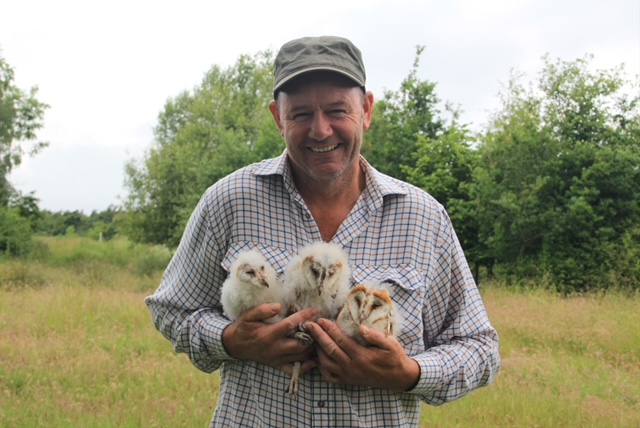 Man in a cap smiles while holding three fluffy baby birds in a grassy field with trees behind him