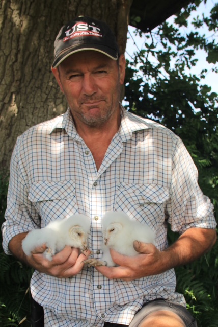 Man in a plaid shirt and black cap holding two white baby owls in his hands outdoors near a tree