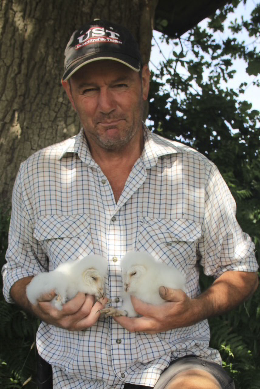 Man in a plaid shirt and black cap holding two fluffy white owls in his hands outdoors near a tree