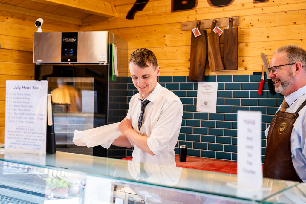 Two men behind a butcher shop counter, one rolling up his sleeves while the other in an apron smiles nearby