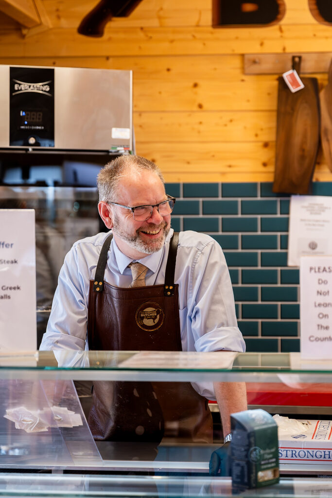 Man in leather apron smiling behind butcher shop counter, with blue tile wall and wooden boards in background
