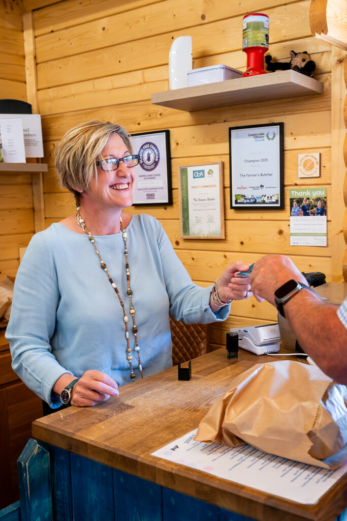 Shop assistant hands a payment card to a customer at a wooden counter, with award certificates on the wall behind
