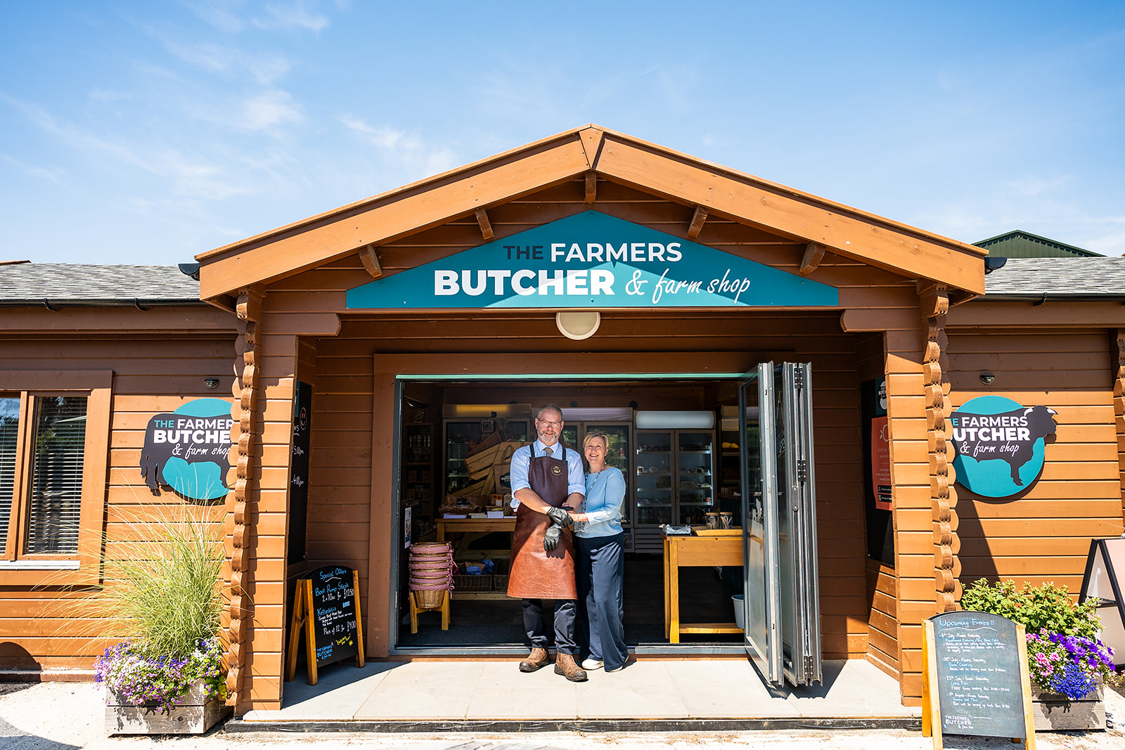Two people stand at the entrance of The Farmers Butcher & farm shop, a wooden storefront with teal signage