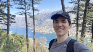 A selfie of a young man with trees behind him and a mountainous background with a lake.