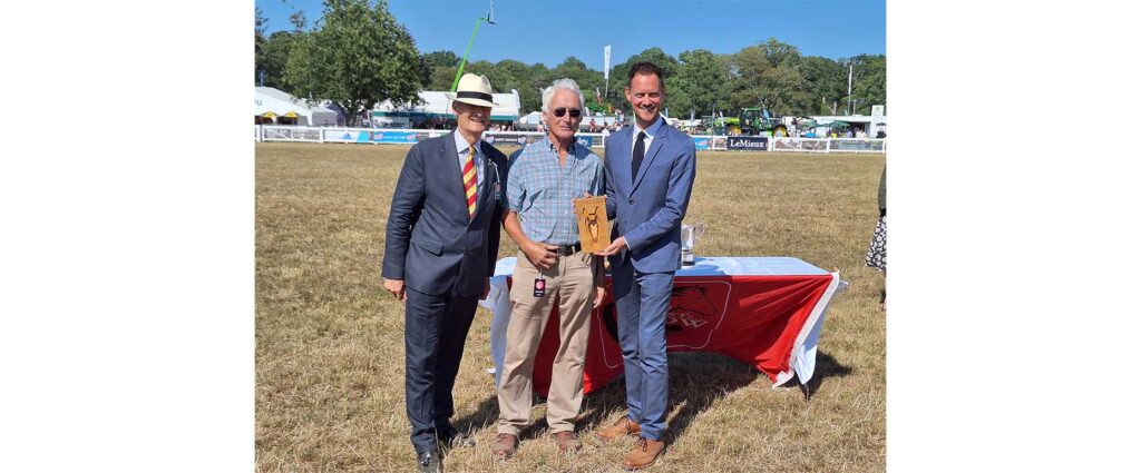 Three men pose at an outdoor show ring, one holding a wooden horse trophy beside a table with silver cups
