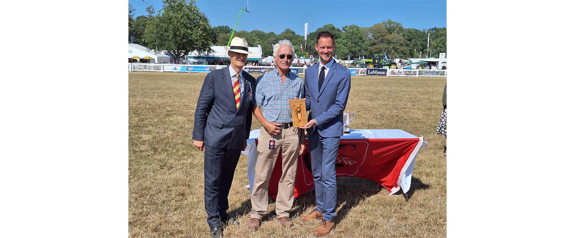 Three men pose at an outdoor show ring, one holding a wooden horse trophy beside a table with silver cups