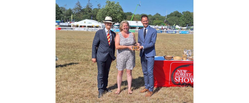 Three people holding an award plaque on a showground field beside a table with trophies and a New Forest and Hampshire County Show banner