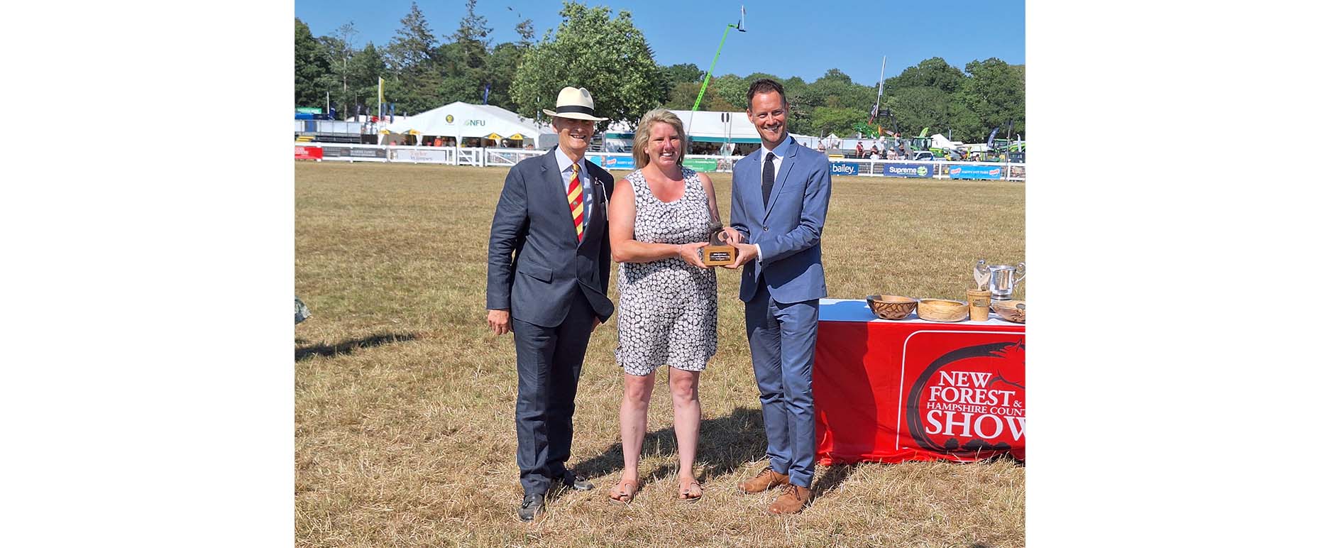 Three people holding an award plaque on a showground field beside a table with trophies and a New Forest and Hampshire County Show banner