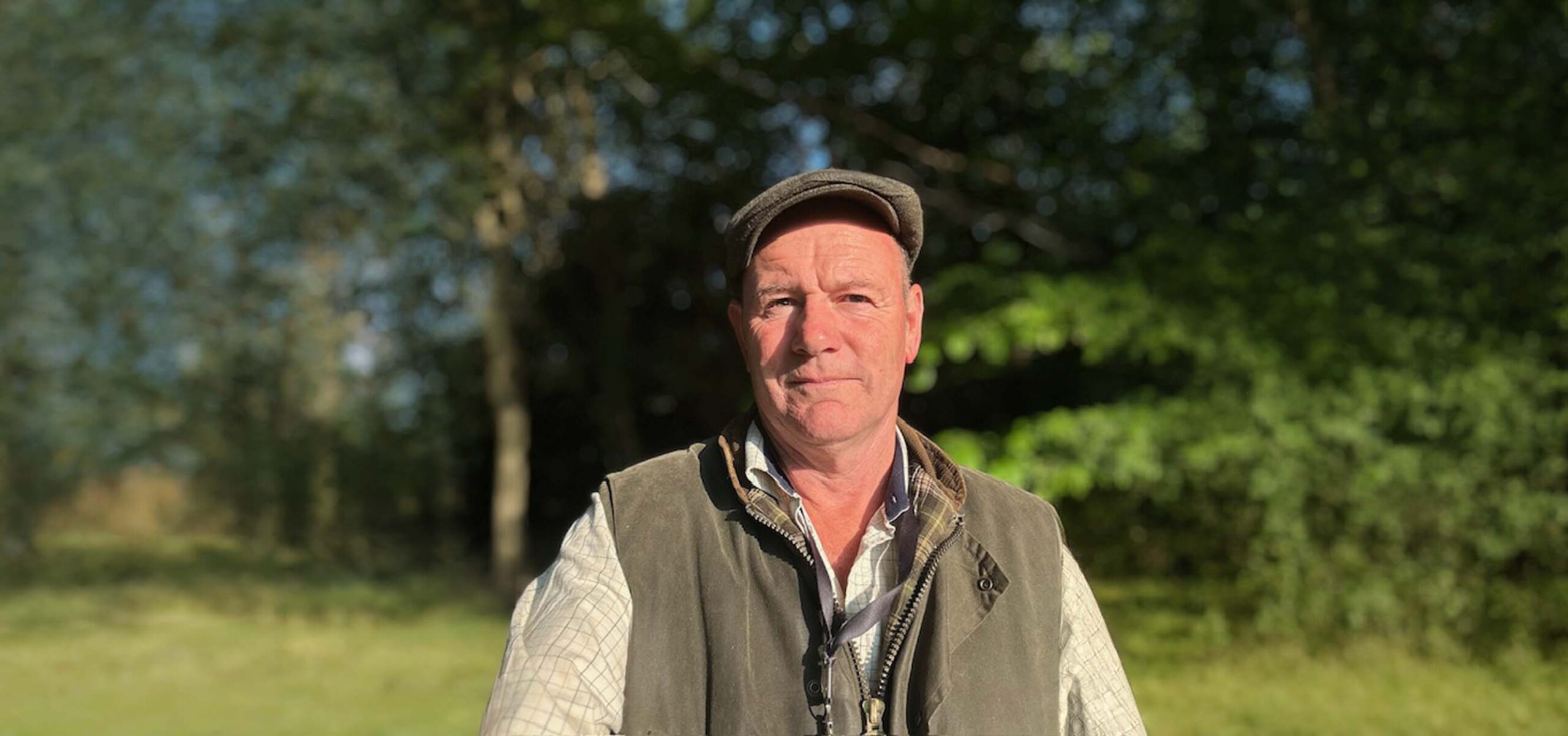 Man wearing flat cap and green vest standing outdoors in sunlight with trees and grass in the background