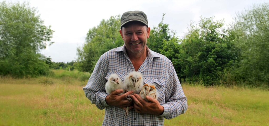 Man in a cap smiles while holding three fluffy baby birds in a grassy field with trees behind him