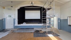 Village hall stage with large projector screen, scaffolding tower at right, and metal poles and floor coverings during renovation