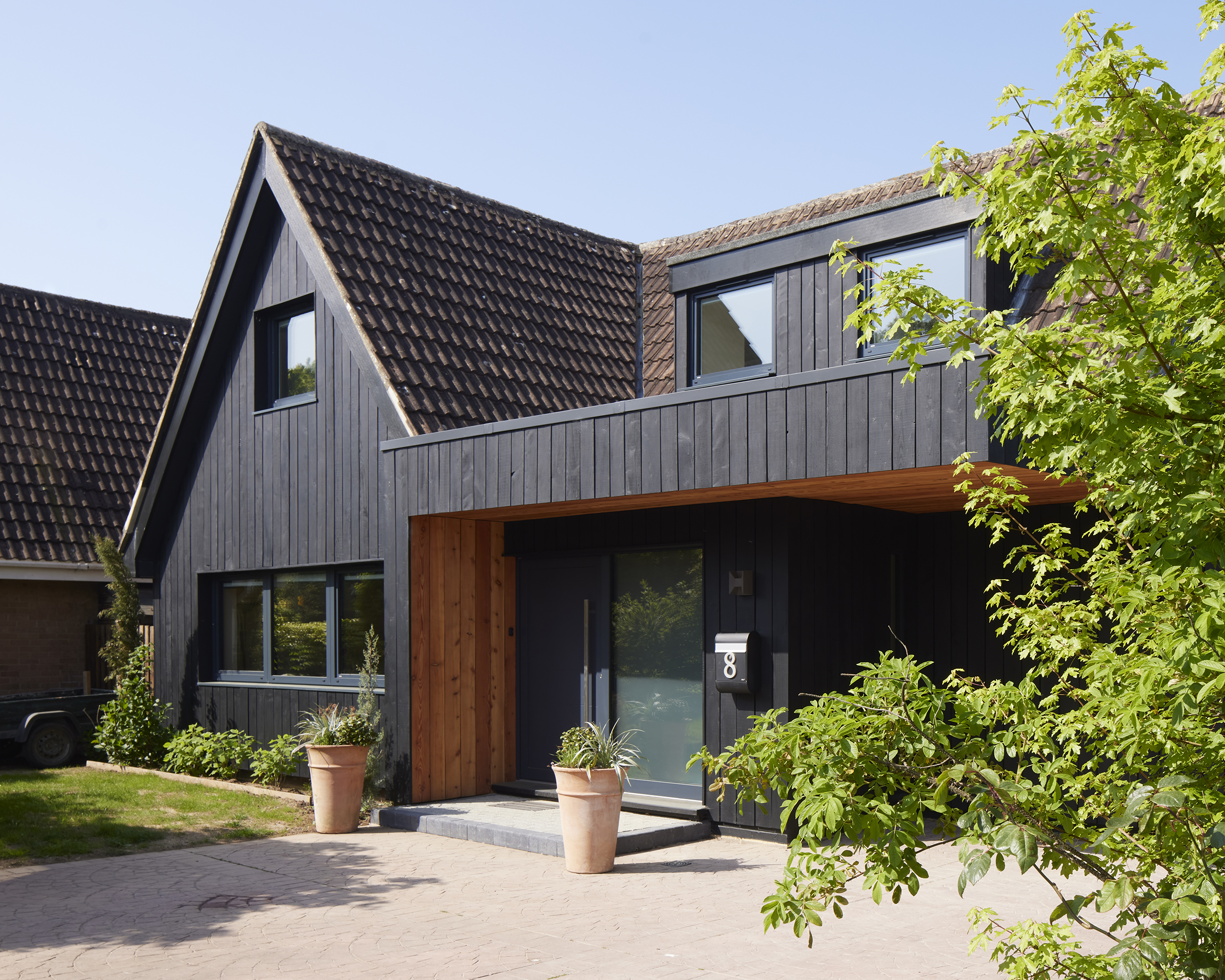 Front of a modern house with dark timber cladding, tiled pitched roof, large windows, and a porch by leafy shrubs and pots