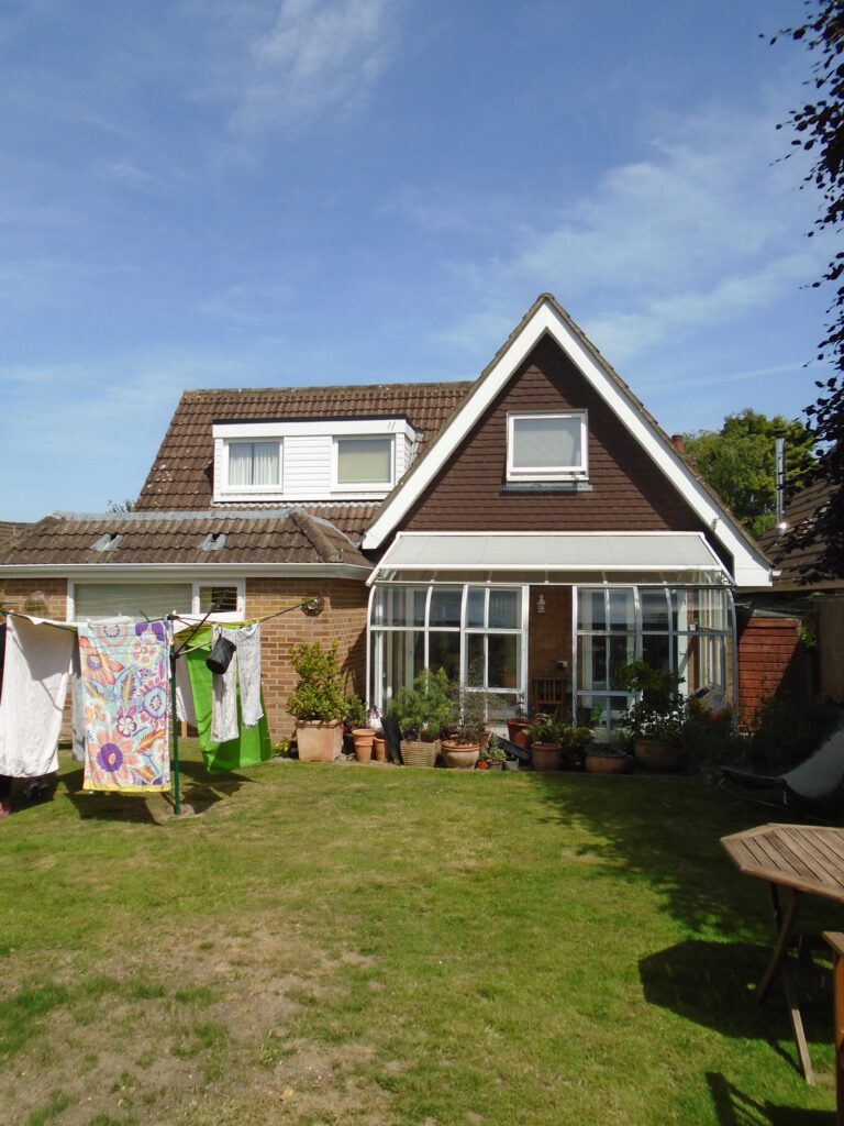 Back view of a house with a conservatory and brown tiled roof, garden lawn with potted plants and laundry on a clothesline under blue sky