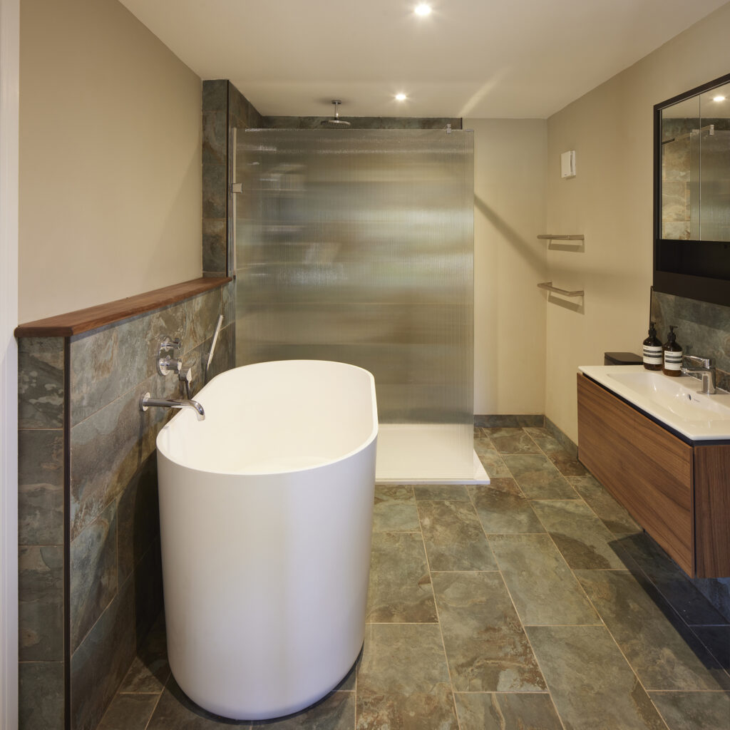 Modern bathroom with freestanding white tub, frosted glass shower screen, slate tile walls and floor, and wood vanity with sink