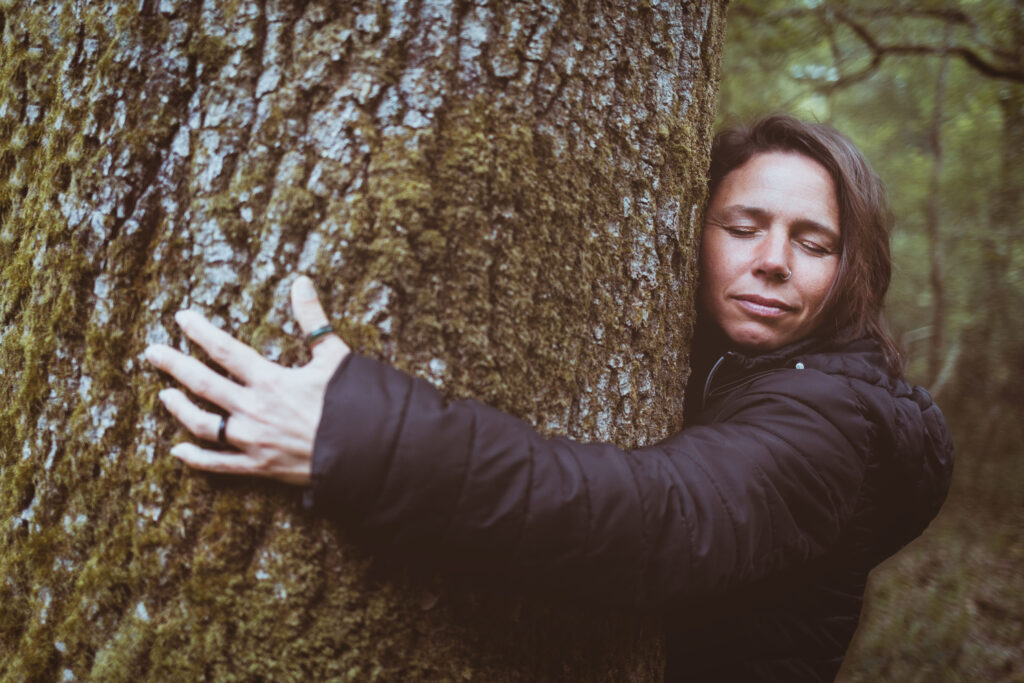 Person with eyes closed hugging a moss-covered tree trunk in a forest, wearing a dark jacket