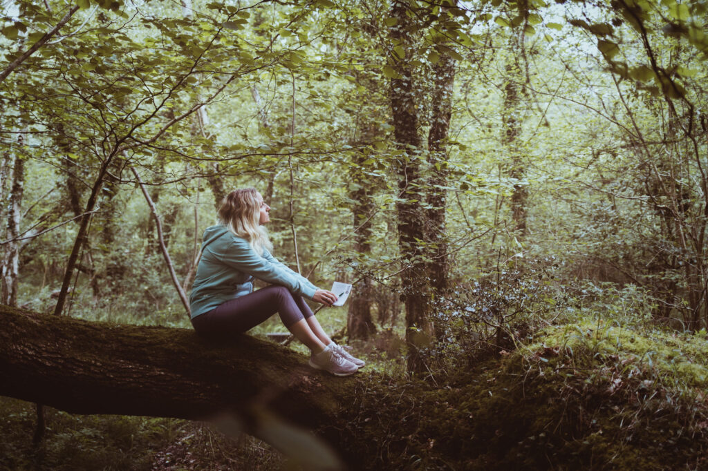 Woman sitting on a mossy fallen tree in a leafy forest, holding a notebook and looking up at the canopy