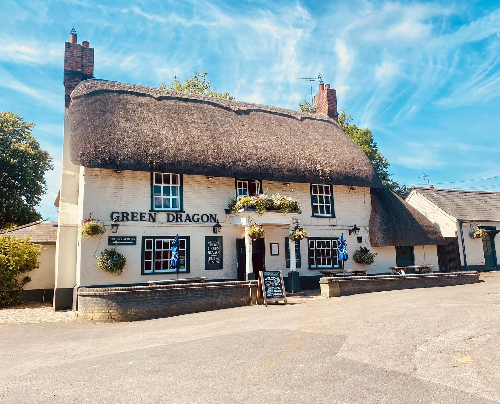 Thatched-roof pub with sign reading Green Dragon, hanging flower baskets, and outdoor tables under a blue sky