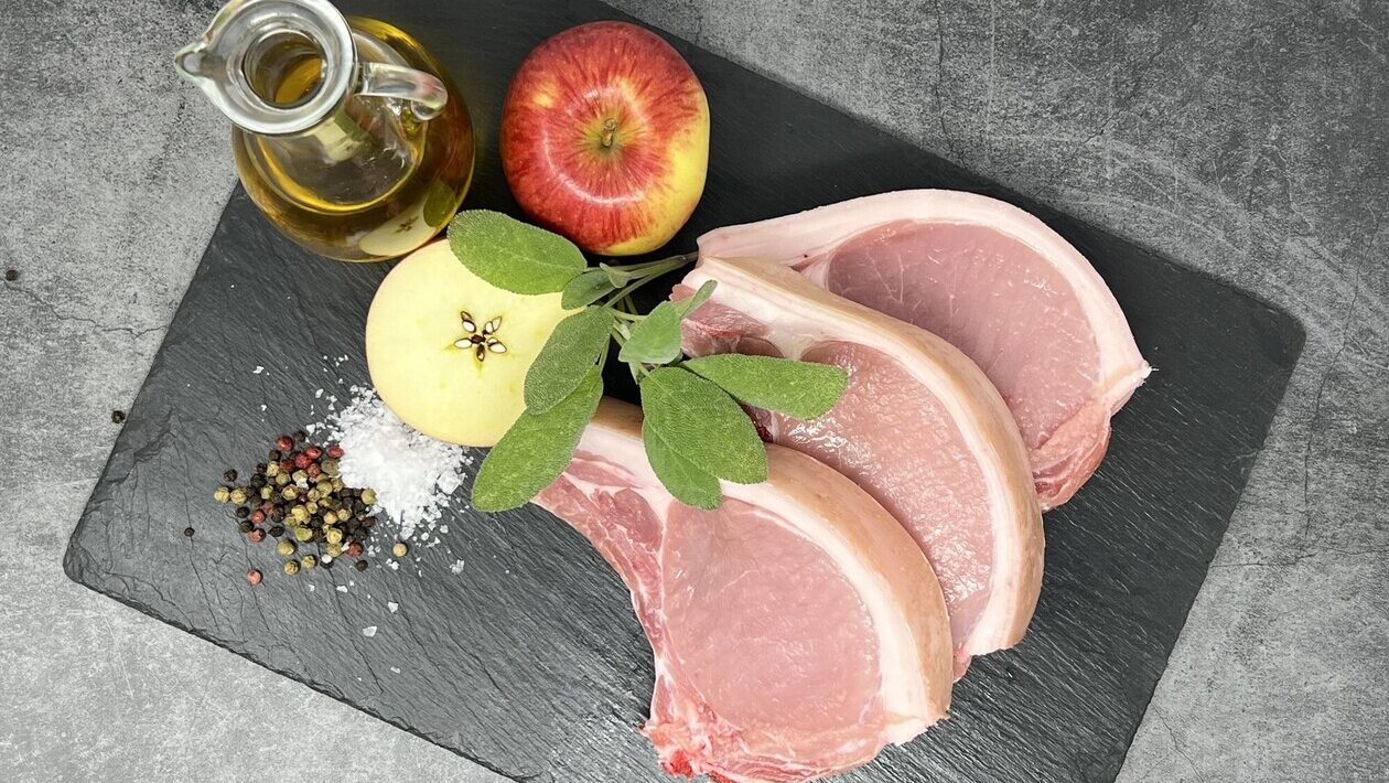 Three raw pork chops on a slate board with sage leaves, apple halves, olive oil, sea salt, and mixed peppercorns