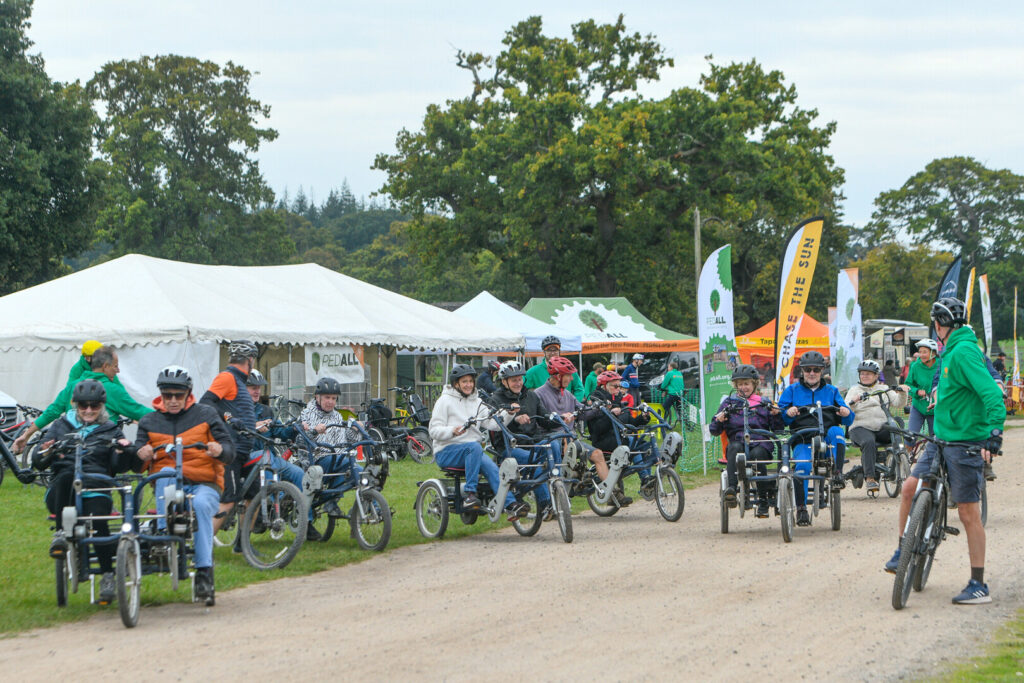 People riding adapted tricycles in a group at an outdoor cycling event, with tents and banners in the background