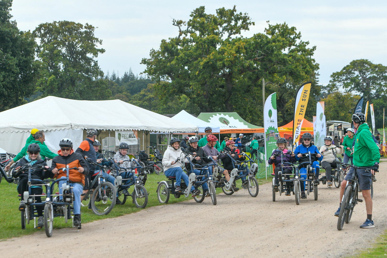 People riding adapted tricycles in a group at an outdoor cycling event, with tents and banners in the background
